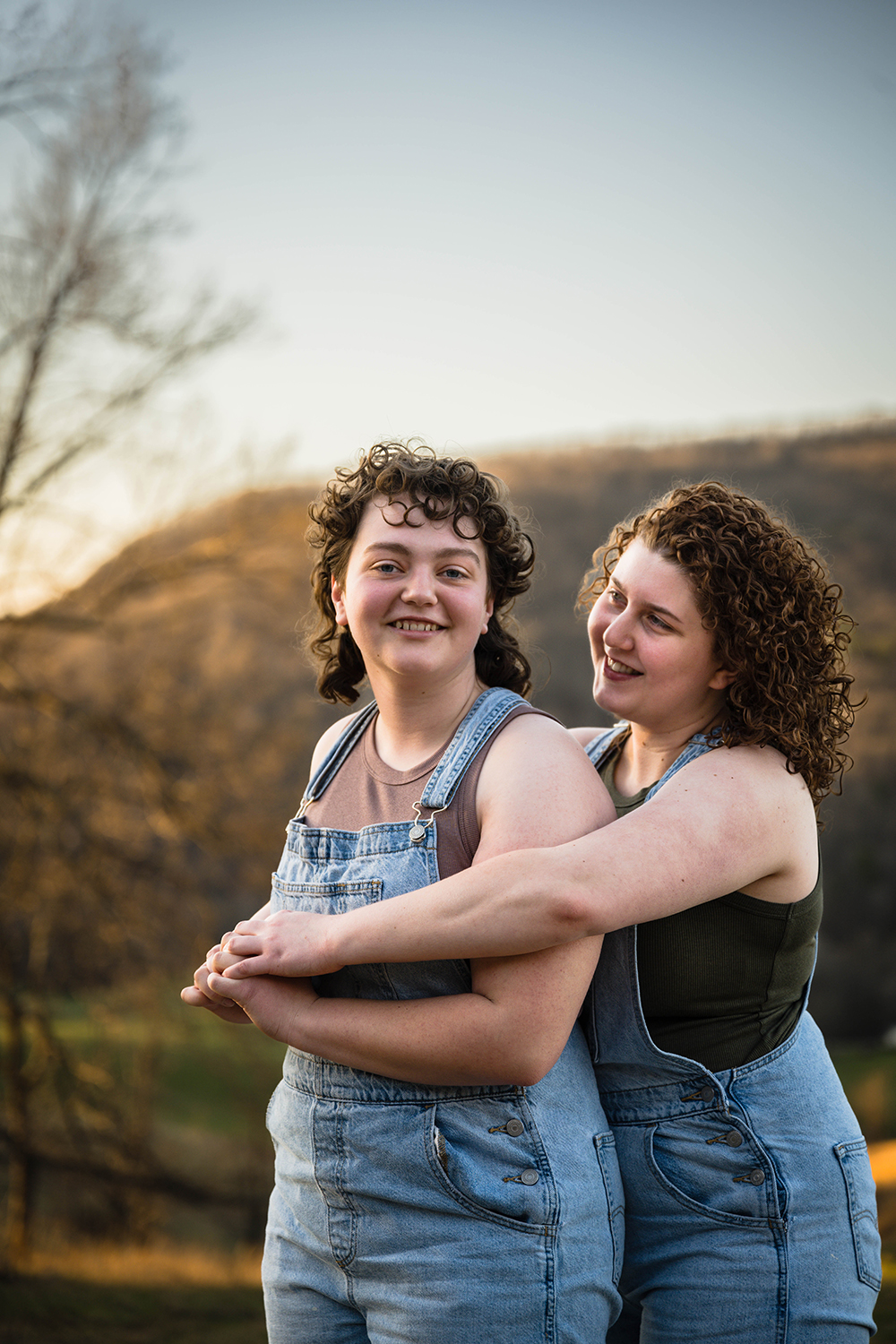 A lesbian woman hugs her partner from behind and looks at her while smiling as her partner looks directly to the camera and smiles.