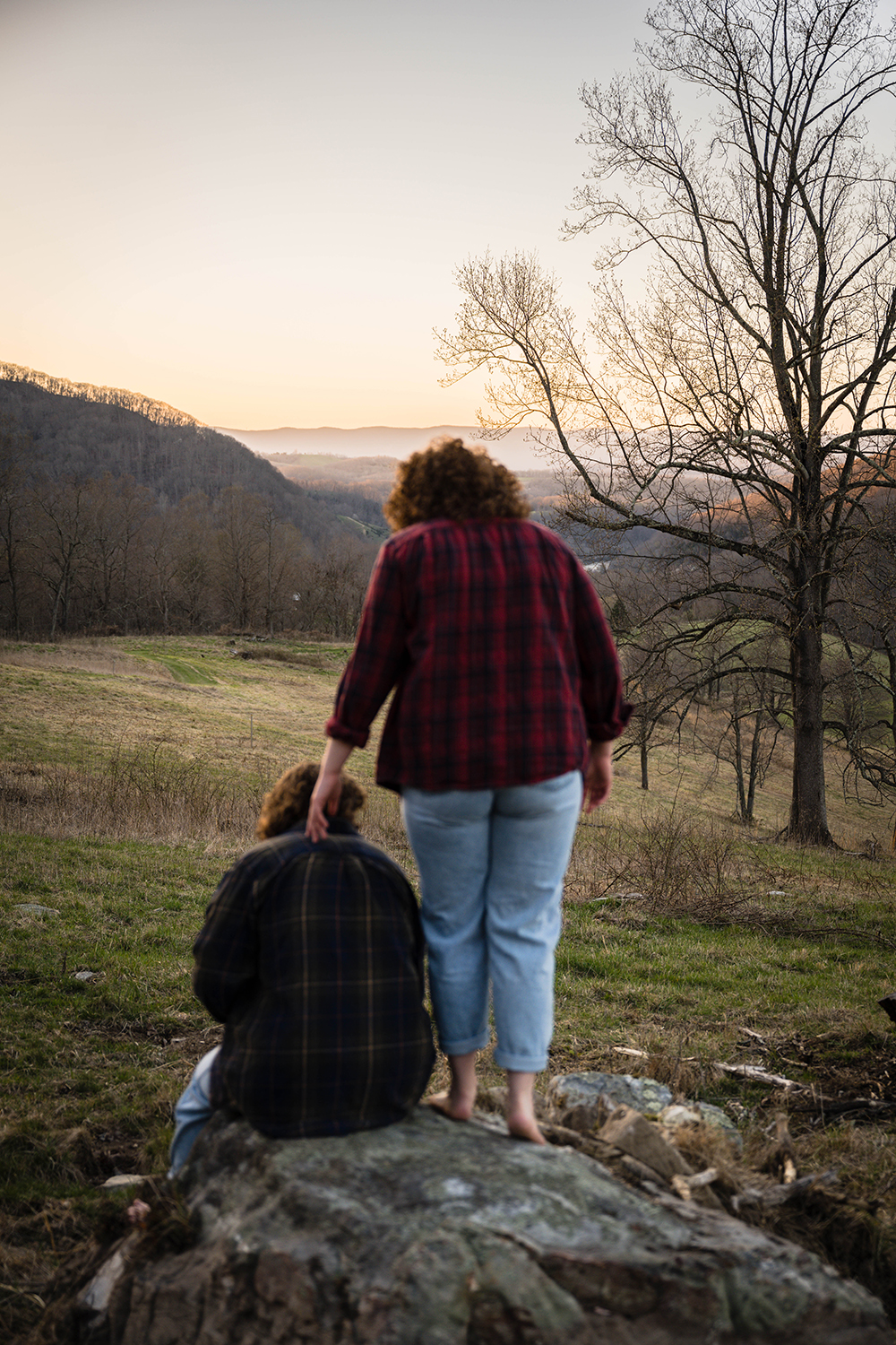 A purposefully out-of-focus queer couples begins to sit down on a large rock while the in-focus sun begins to set behind the Blue Ridge Mountains, which now look more purple.