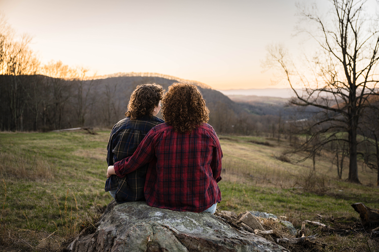 A queer couple sits on a large rock together and have their backs face the camera as they enjoy looking at the sunset together during their anniversary portrait session.