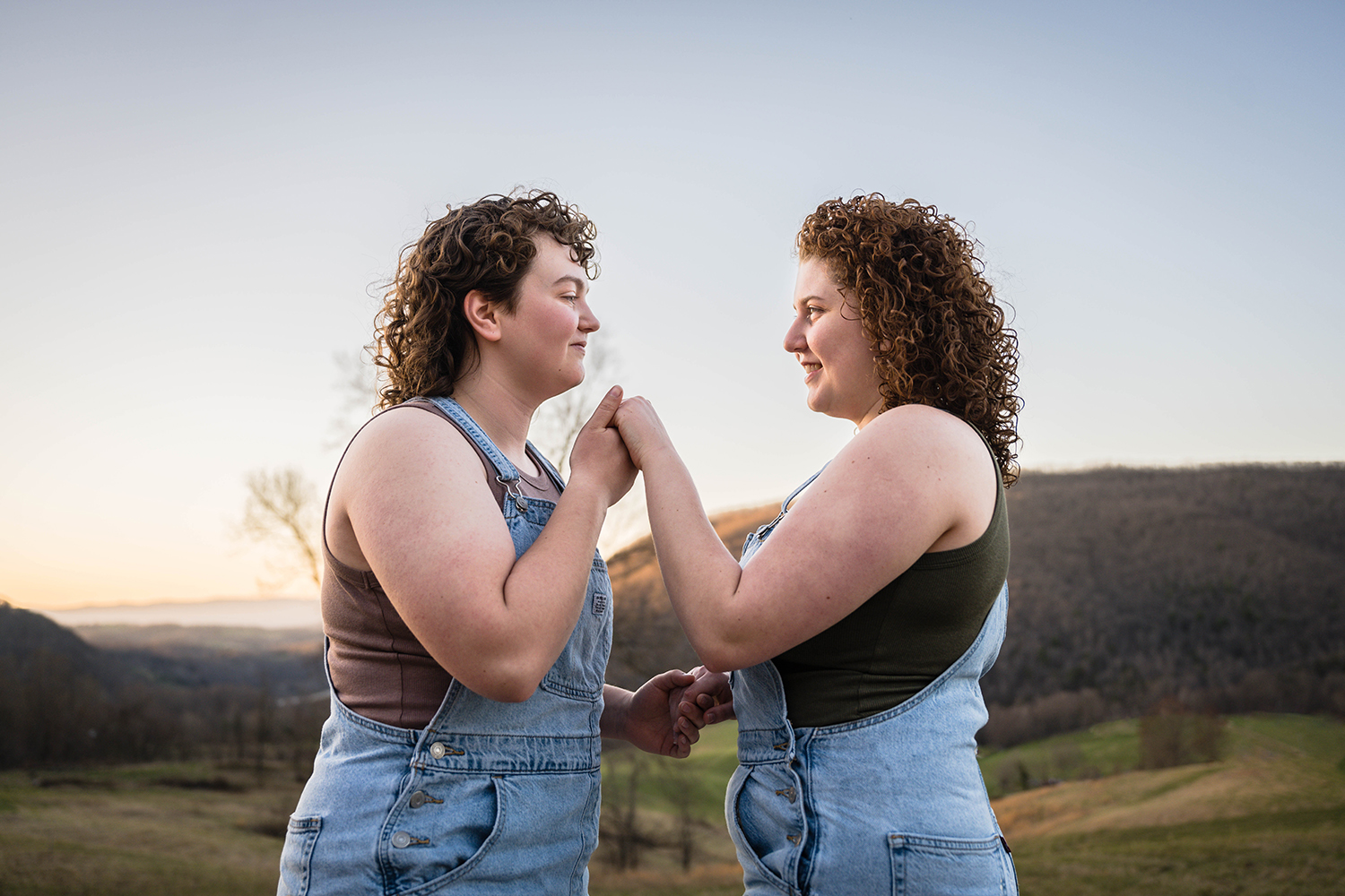 A queer person lifts their partner's hand towards their lips as to prepare to give their hand a kiss while their partner looks knowingly. 