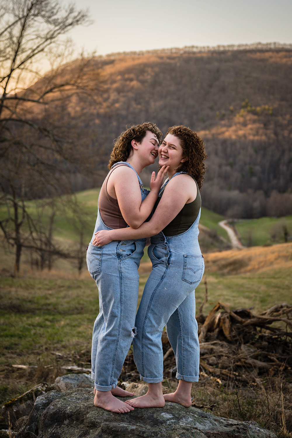 A queer individual holds the face of their partner and smiles as they go to kiss their cheek while their partner hangs onto them and smiles while looking away from the camera.