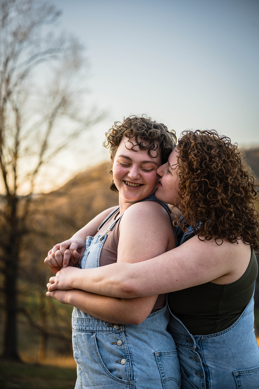 A lesbian woman hugs her significant other from behind and kisses her cheek while her partner leans into her and smiles while looking down at their shoulder.