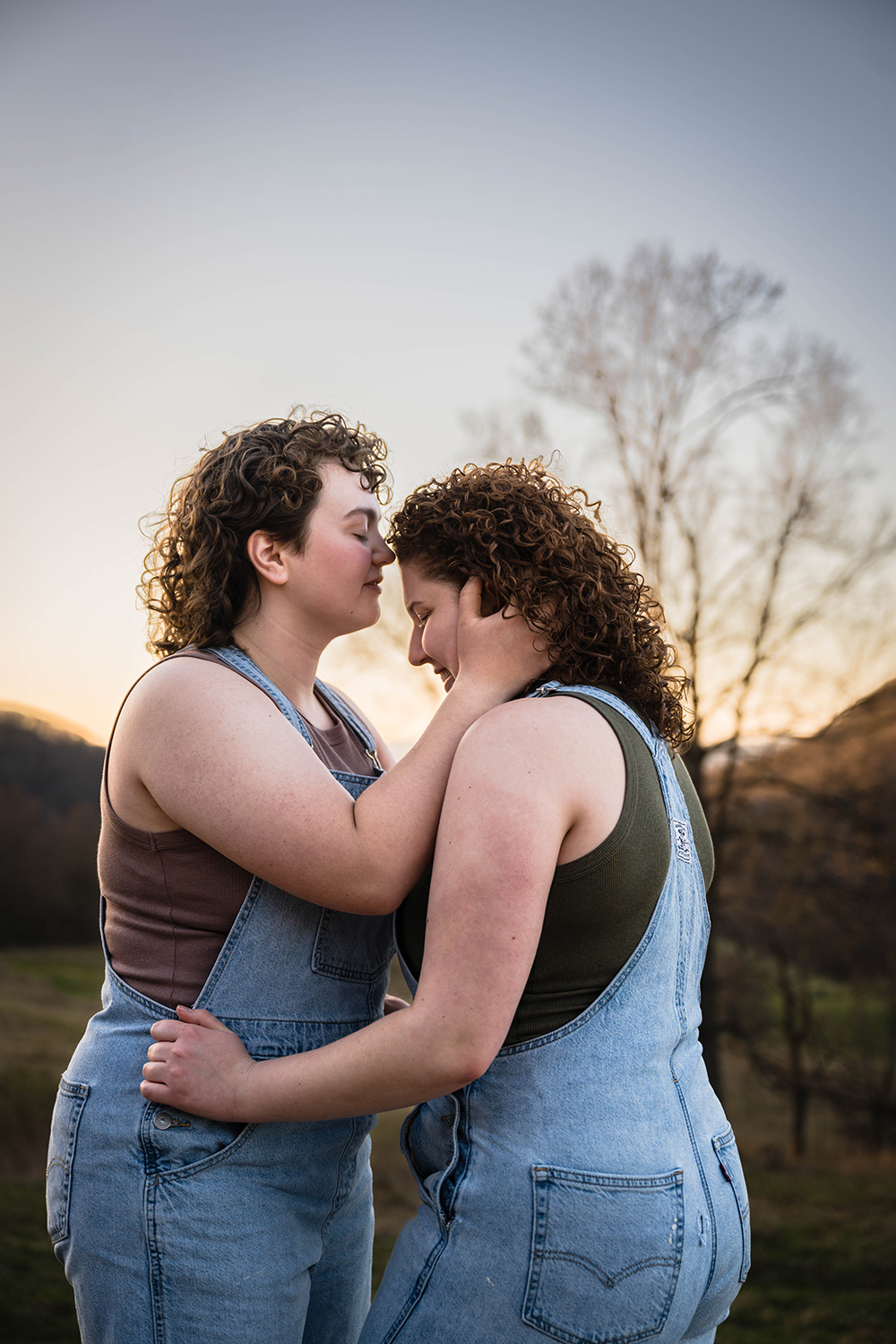 A lesbian woman holds onto her partner's hips as her partner holds her head and brings it close to their mouth to prepare to give a forehead kiss.