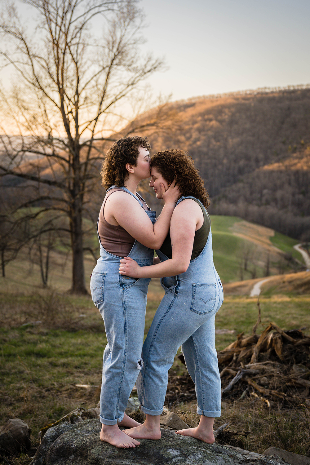 A lesbian woman holds onto her partner's hips as her partner gives her a kiss on the forehead.