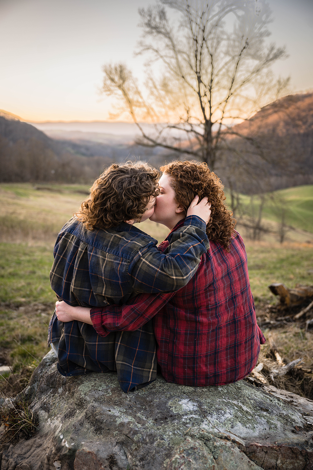 A queer couple sits on a large rock together and have their backs face the camera as they give each other a kiss during their anniversary portrait session.