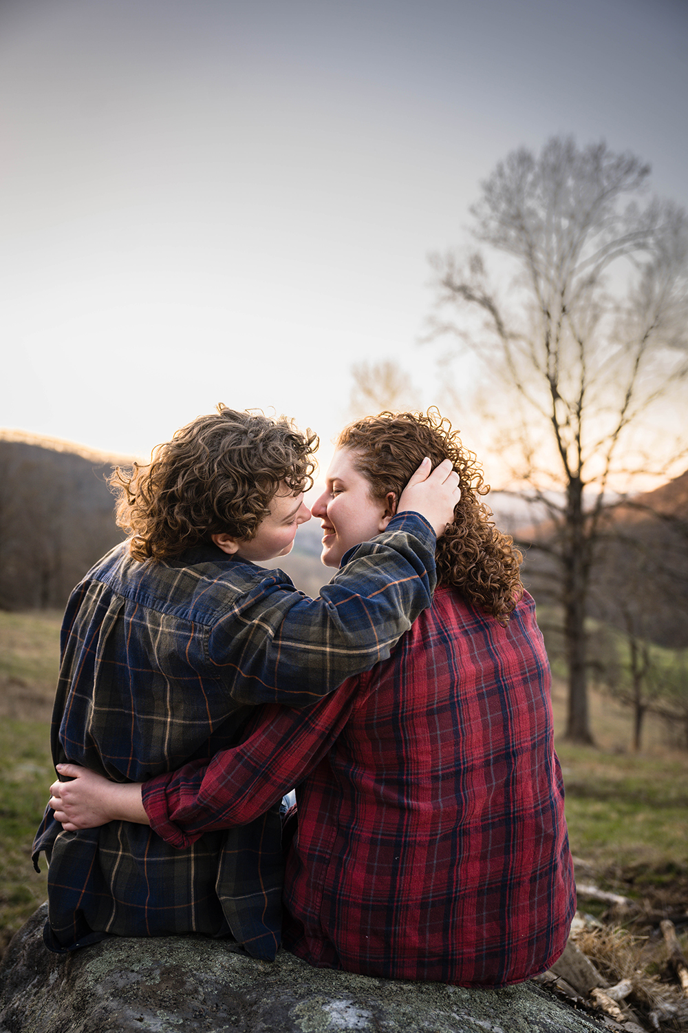 A queer couple sits on a large rock together and have their backs face the camera as they go in for a kiss during their anniversary portrait session.