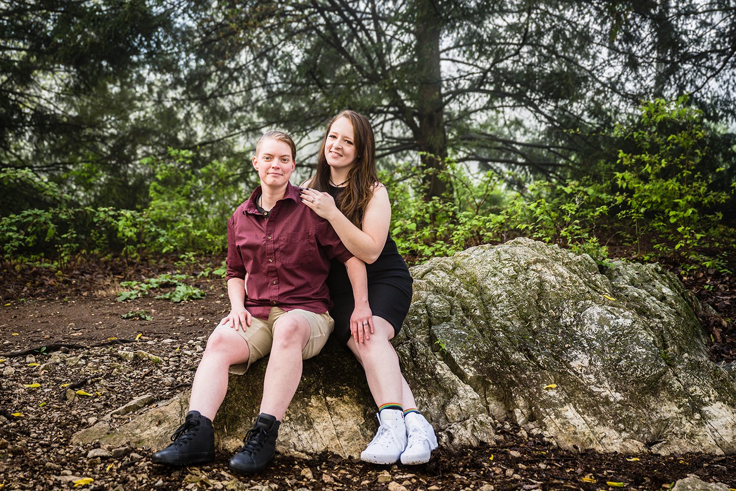 An lgbtq+ couple sits on a large rock located at the summit of Mill Mountain for their engagement session in Virginia.