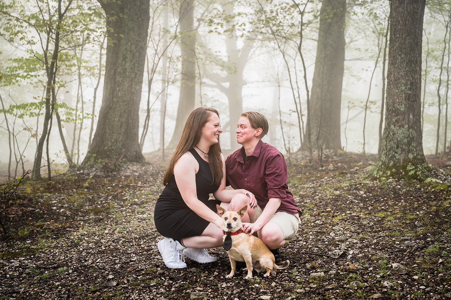 An lgbtq couple smile at one another and hold onto their dog for Roanoke Star engagement session.