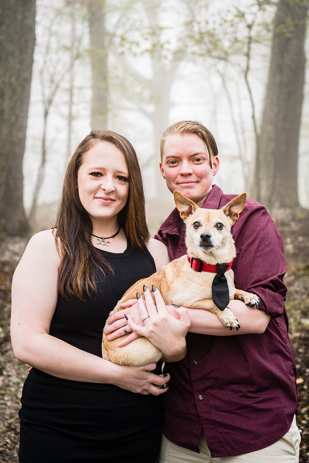 An lgbtq couple smile for the camera and hold onto their dog during their engagement session.