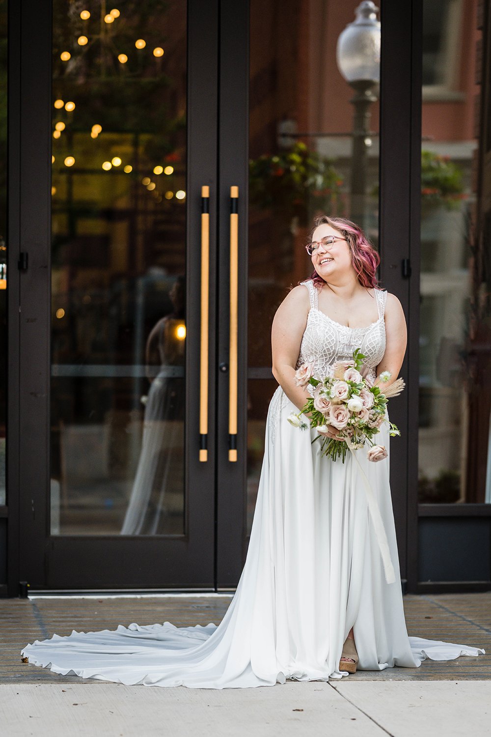 A wedding bride stands in front of the Fire Station One holds their wedding bouquet and smiles looking away from the camera.