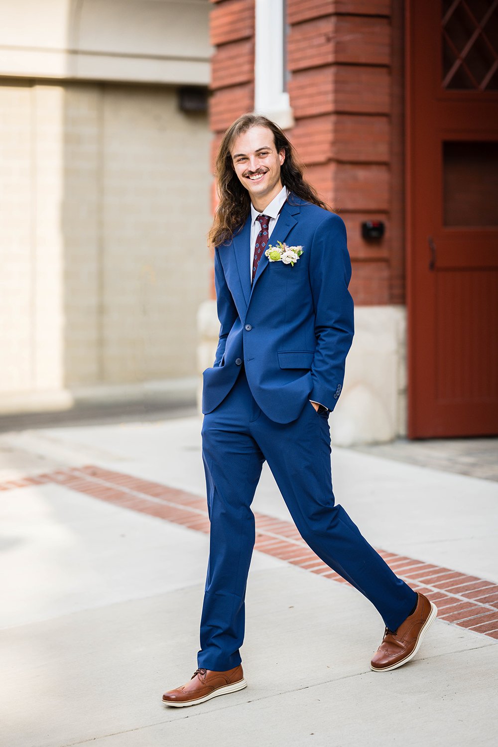 A groom walks with his hands in his pockets in front of the Fire Station One Hotel and smiles away from the camera.