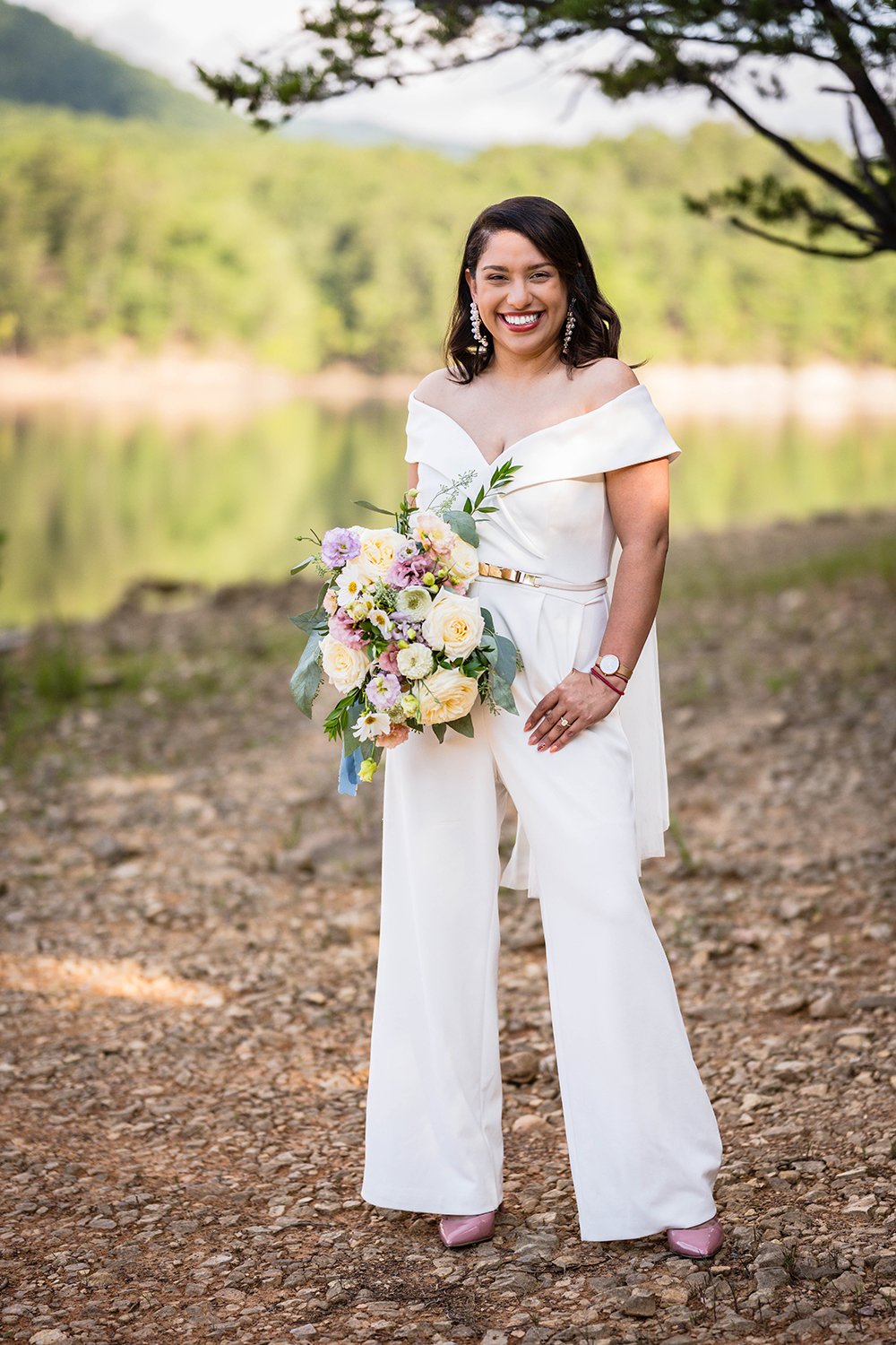 A smiling bride sporting a beautiful and bold bridal jumpsuit holds her wedding bouquet during her elopement at Carvin’s Cove in Roanoke, Virginia.