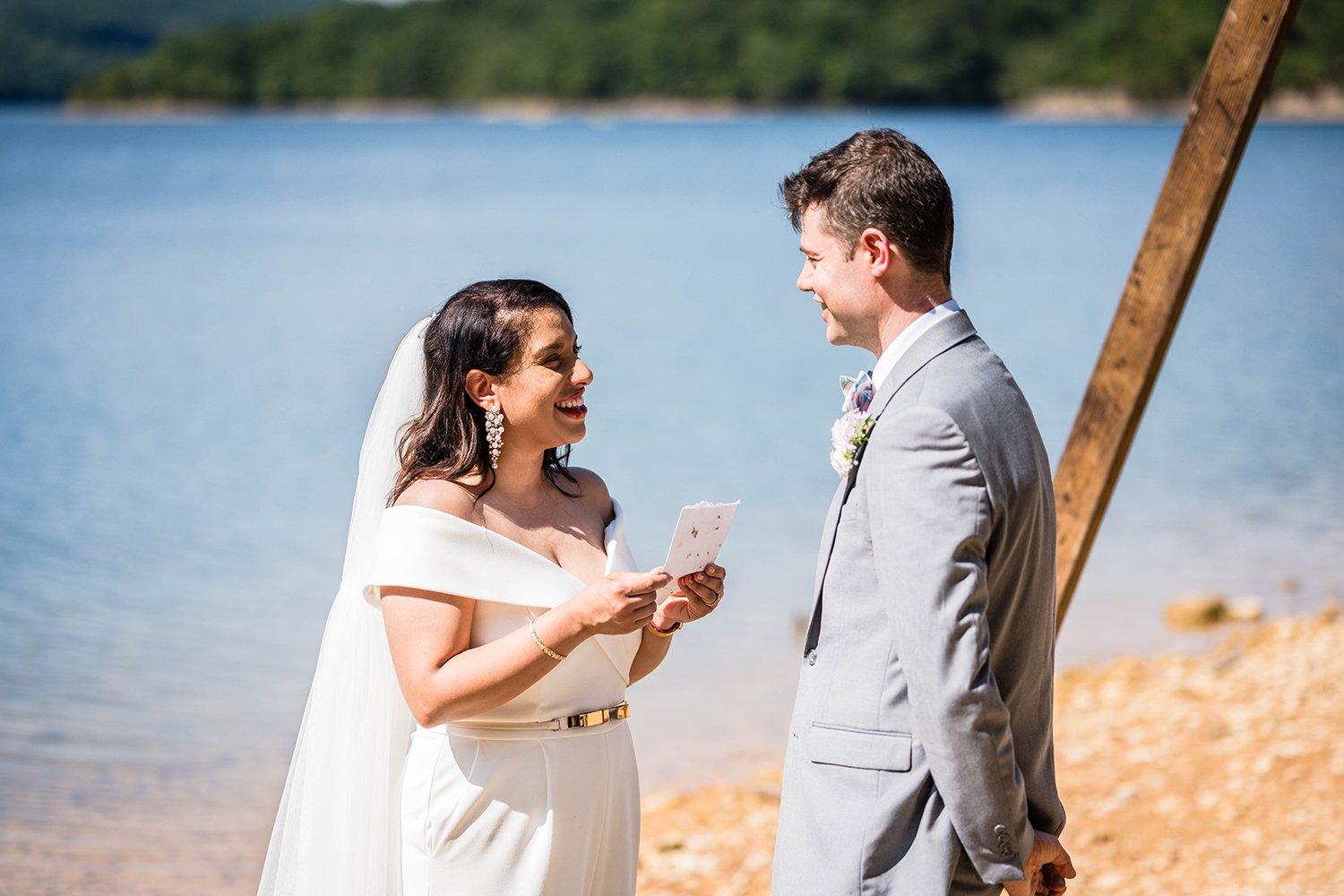 A bride reads her vows along the shoreline of Carvin’s Cove in front of a wooden arch and the groom smiles at her during their elopement.
