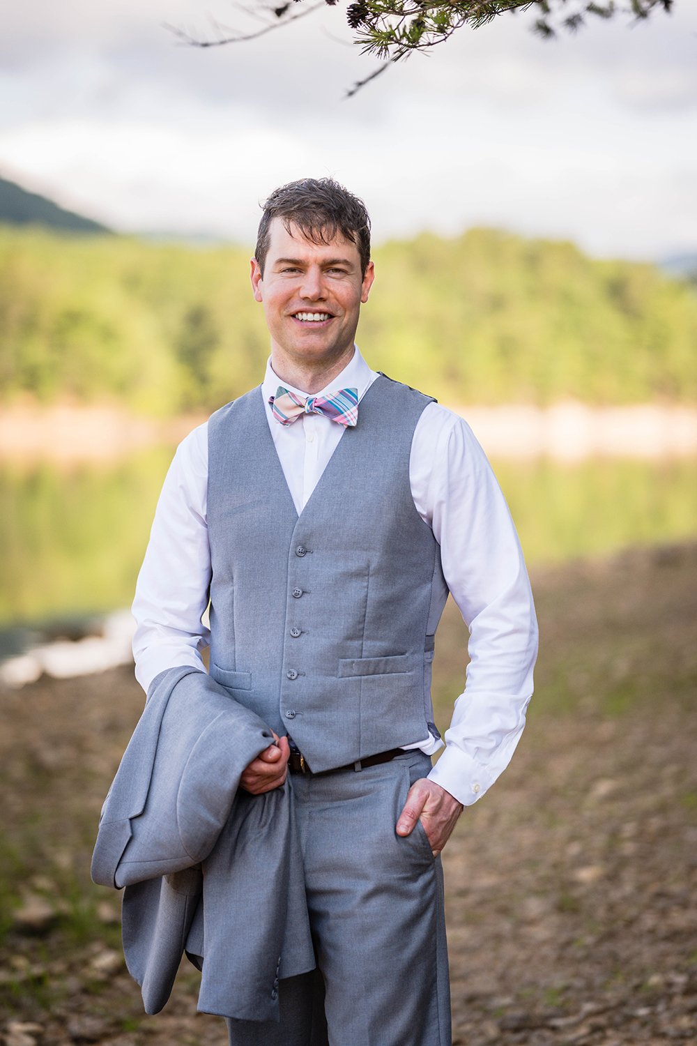 A groom stands holding his suit jacket in one arm and with his other hand in his pocket and smiles for a photo during his elopement at Carvin’s Cove in Southwest Virginia.