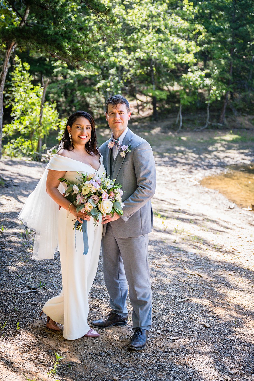A newlywed couple stands underneath a canopy of trees and next to the shoreline for a photo together during their elopement.