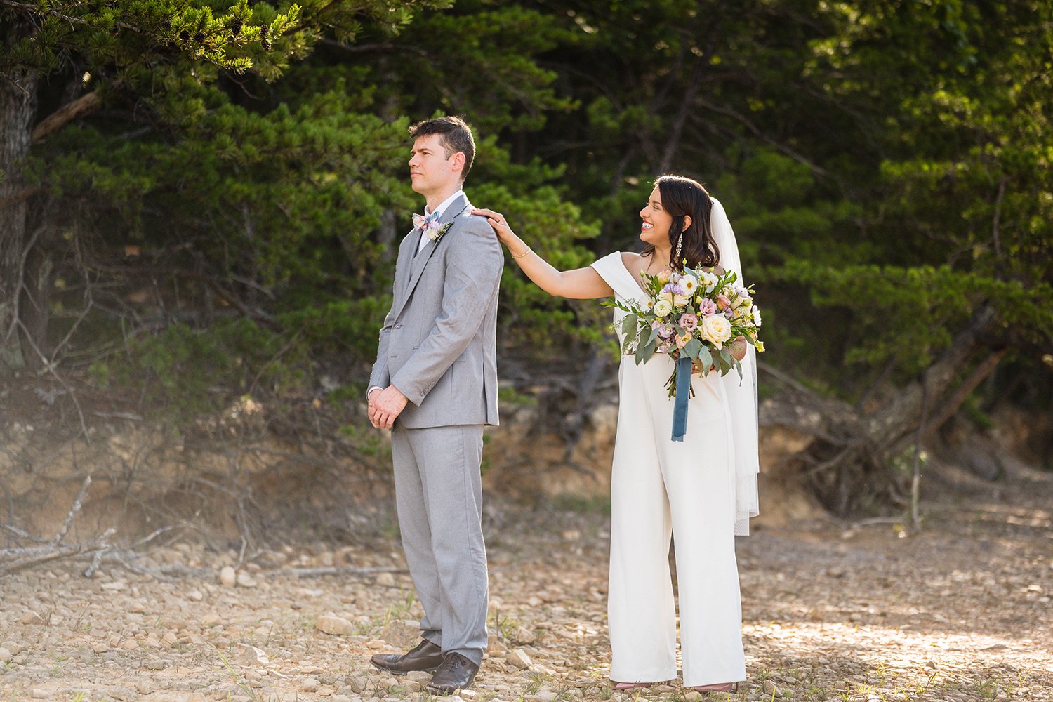 A marrier wearing a white jumpsuit and veil and holding a wedding bouquet taps their partner on the shoulder during their first look at Carvin’s Cove.