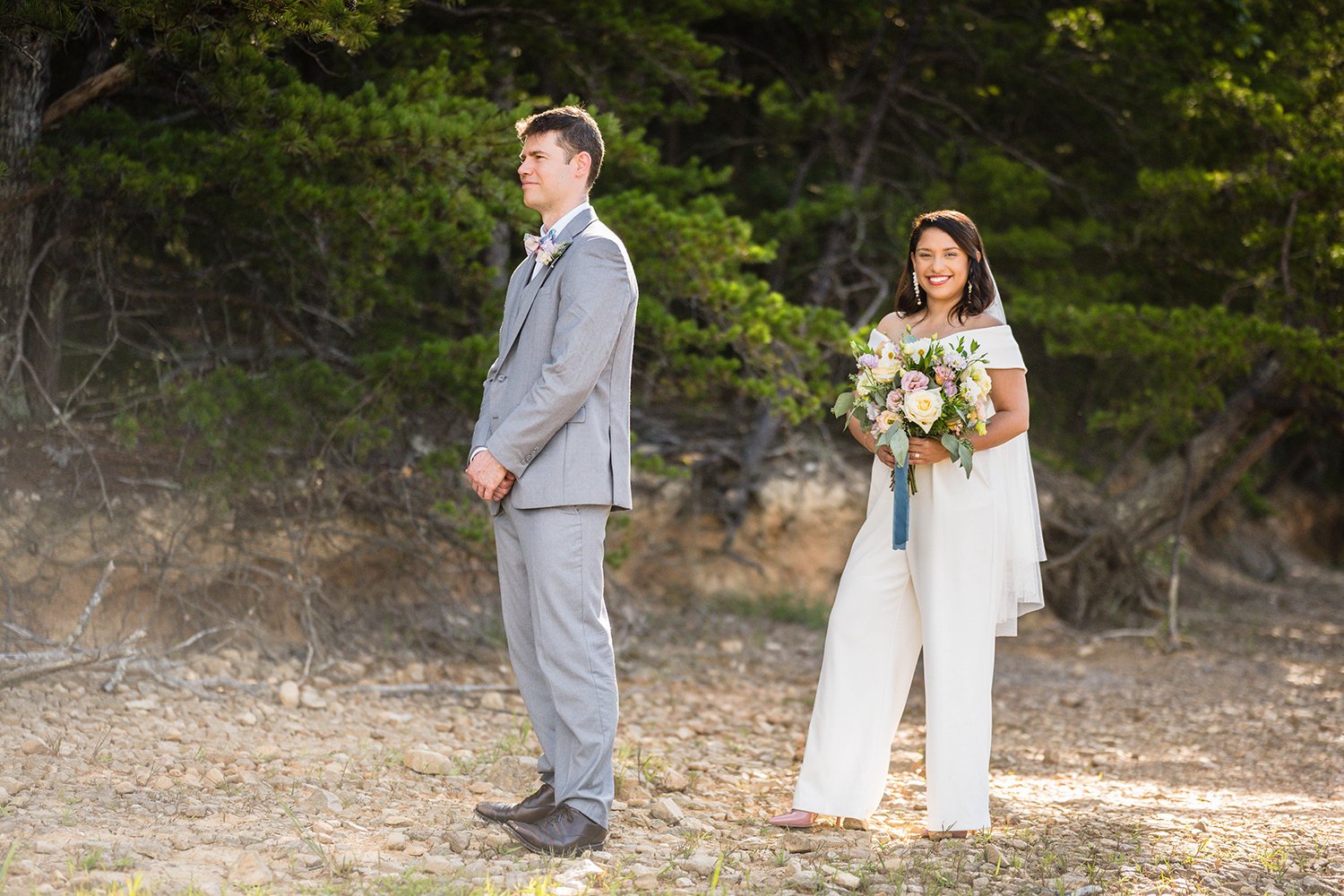 A groom stands facing away from his bride who looks towards the camera smiling and holding her wedding bouquet as she waits for their first look during their elopement in Virginia at Carvin’s Cove.