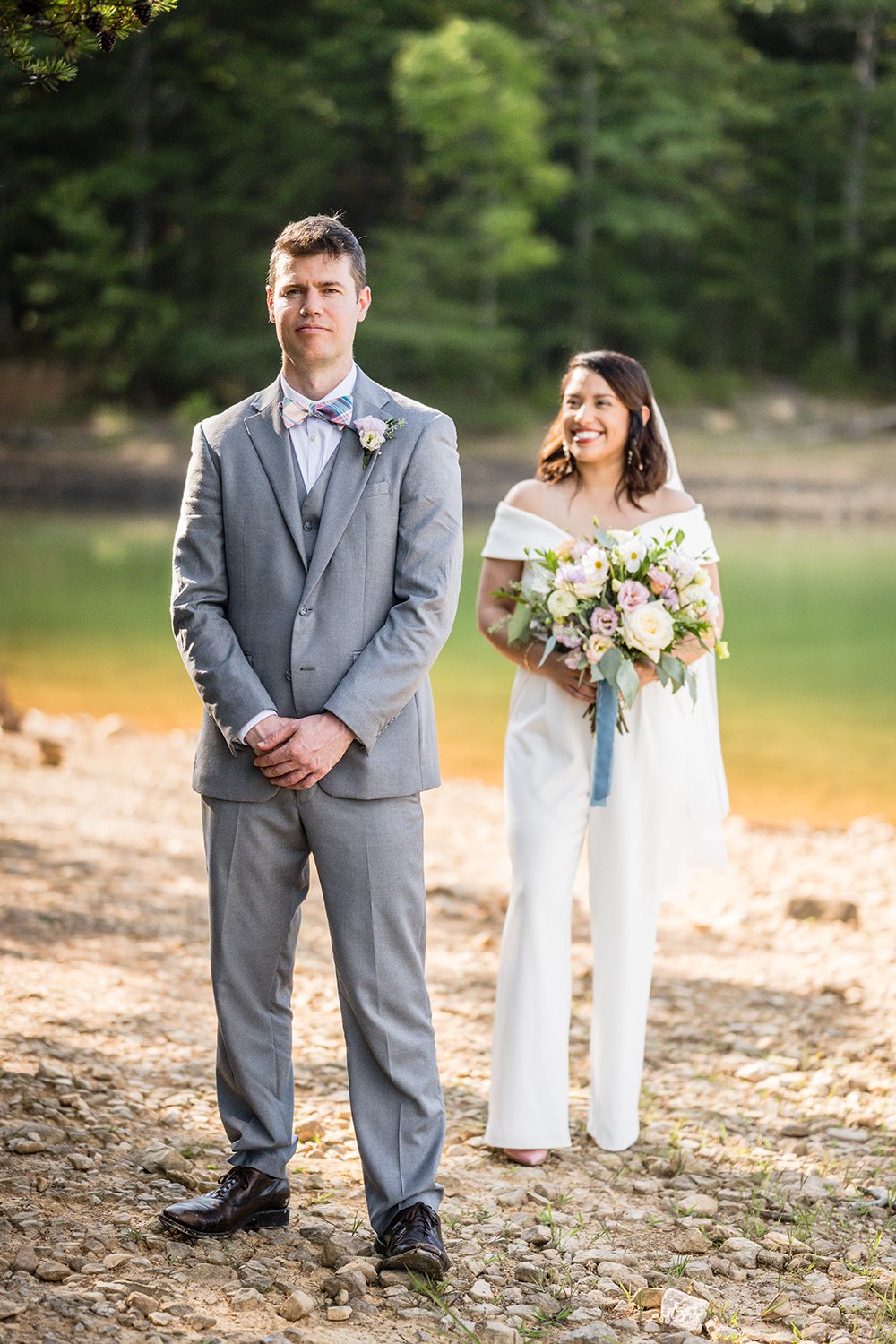 A groom stands facing away from his bride who looks and smiles at the back of his head holding her wedding bouquet as she waits for their first look.