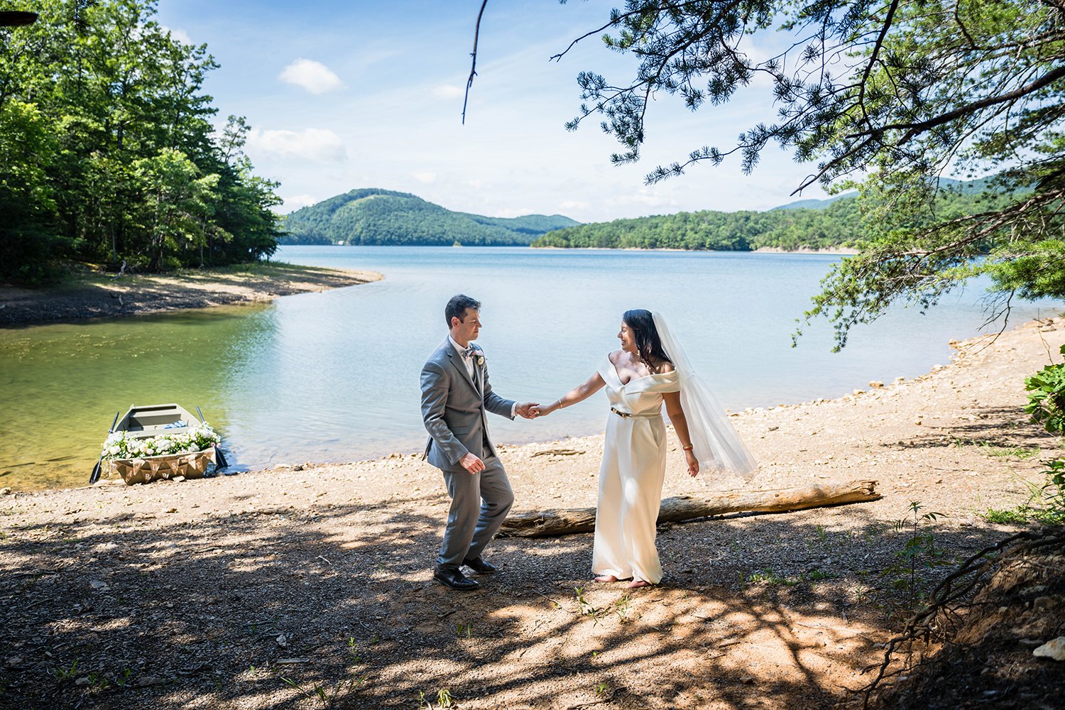 A bride and groom take their first dance on the sandy and rocky shores of Carvin's Cove near Roanoke, Virginia with their rowboat adorned with flowers in the background.