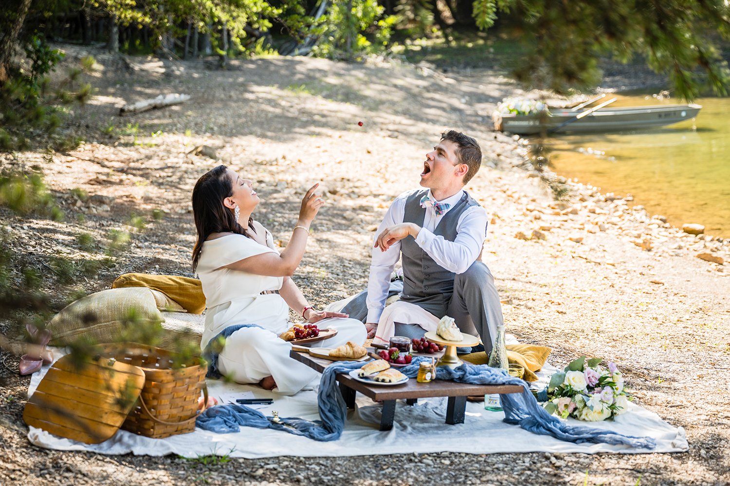 A bride throws a grape in the air as her groom attempts to catch it with his mouth wide open during their brunch picnic session of their elopement timeline.
