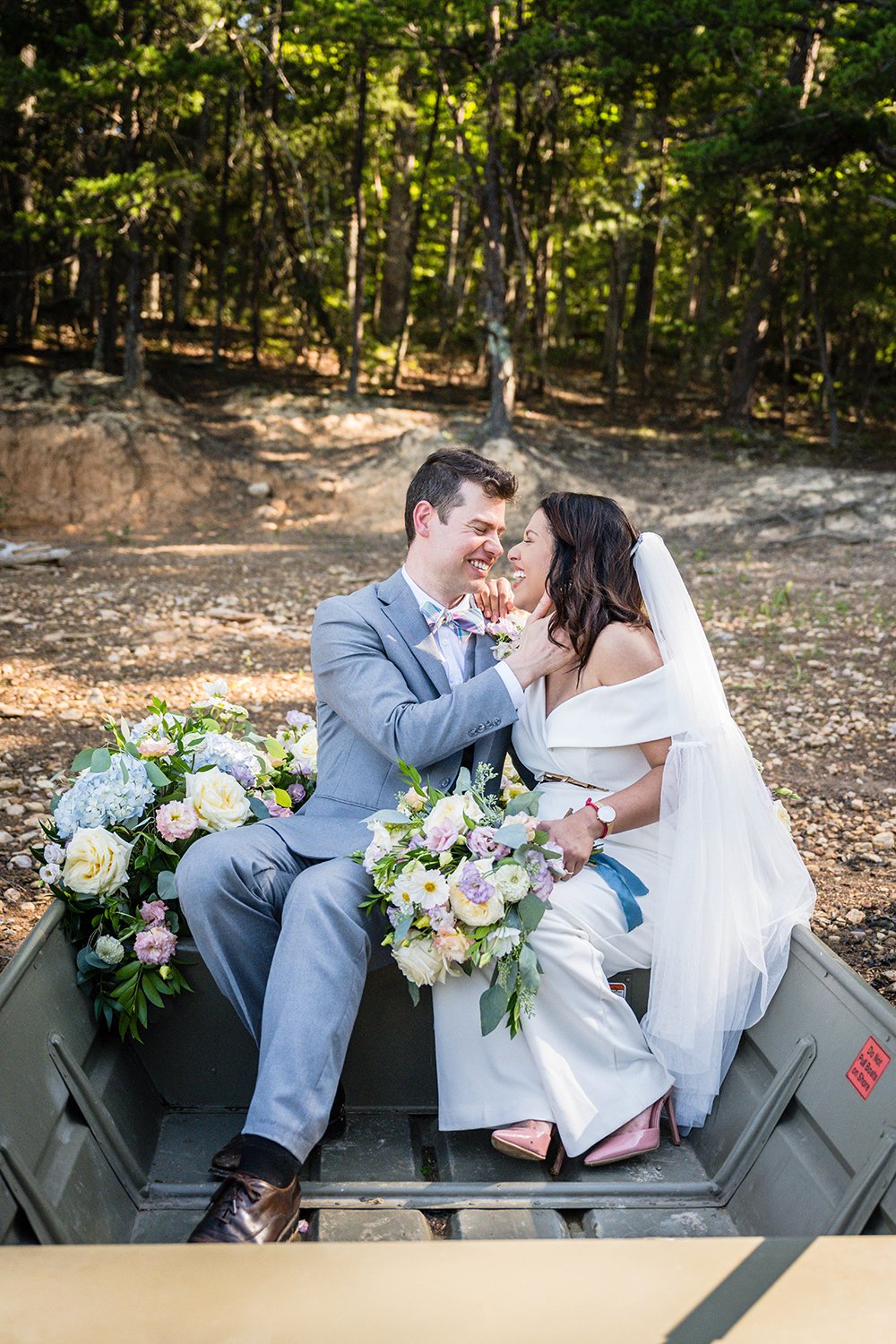 A newlywed couple sits together and holds onto one another laughing in a rowboat adorned with pastel flowers on the shores of Carvin's Cove.