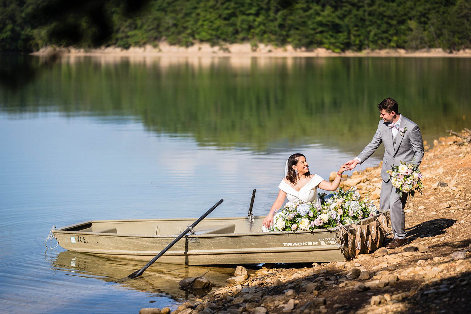 A groom helps his bride into a rowboat adorned with flowers and with a banner that says "just married" while holding a bouquet of flowers on their elopement day at Carvin's Cove.
