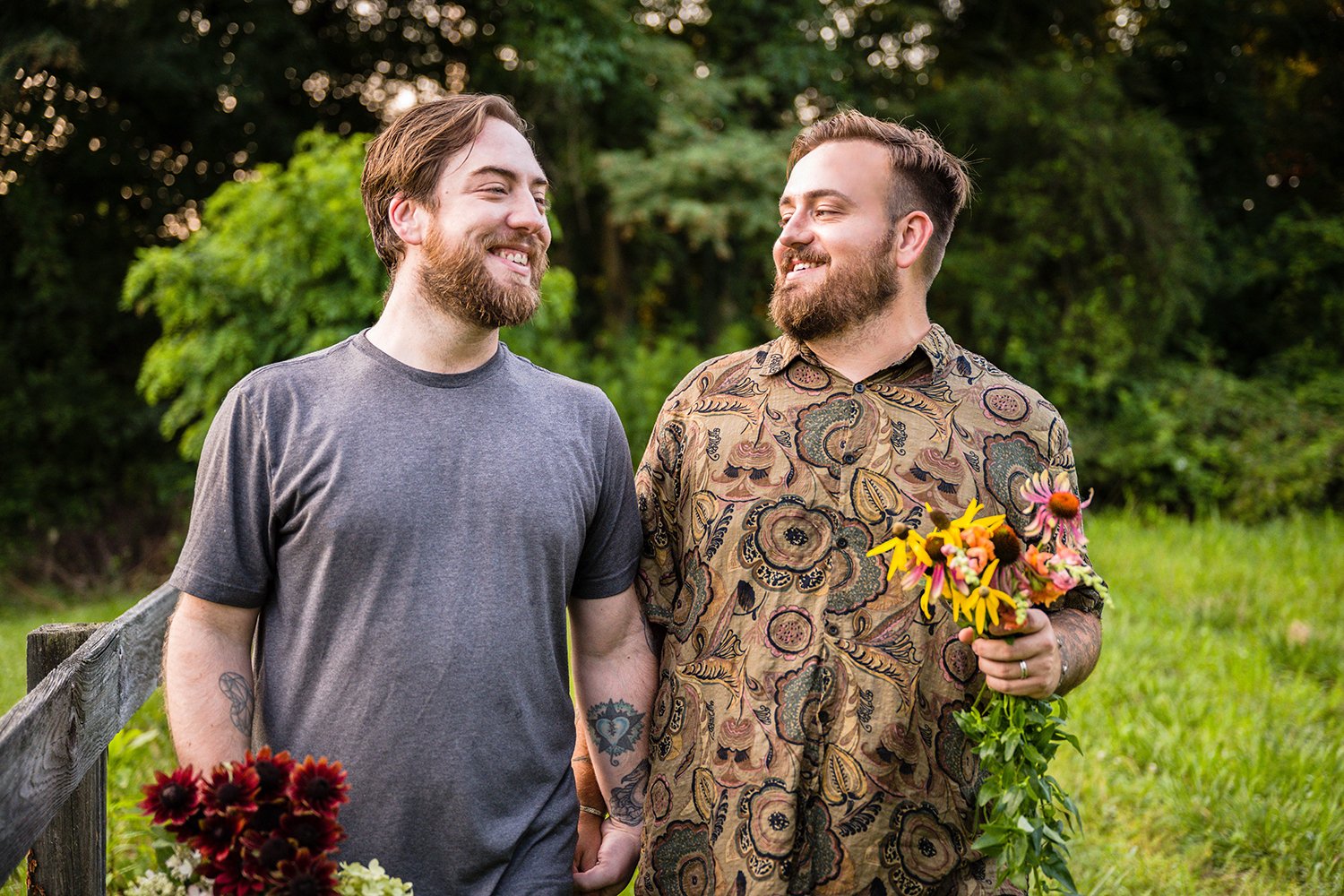 A queer couple holding flowers and one another's hands smiles towards one another on their farm, Fae Cottage Flower Farm.