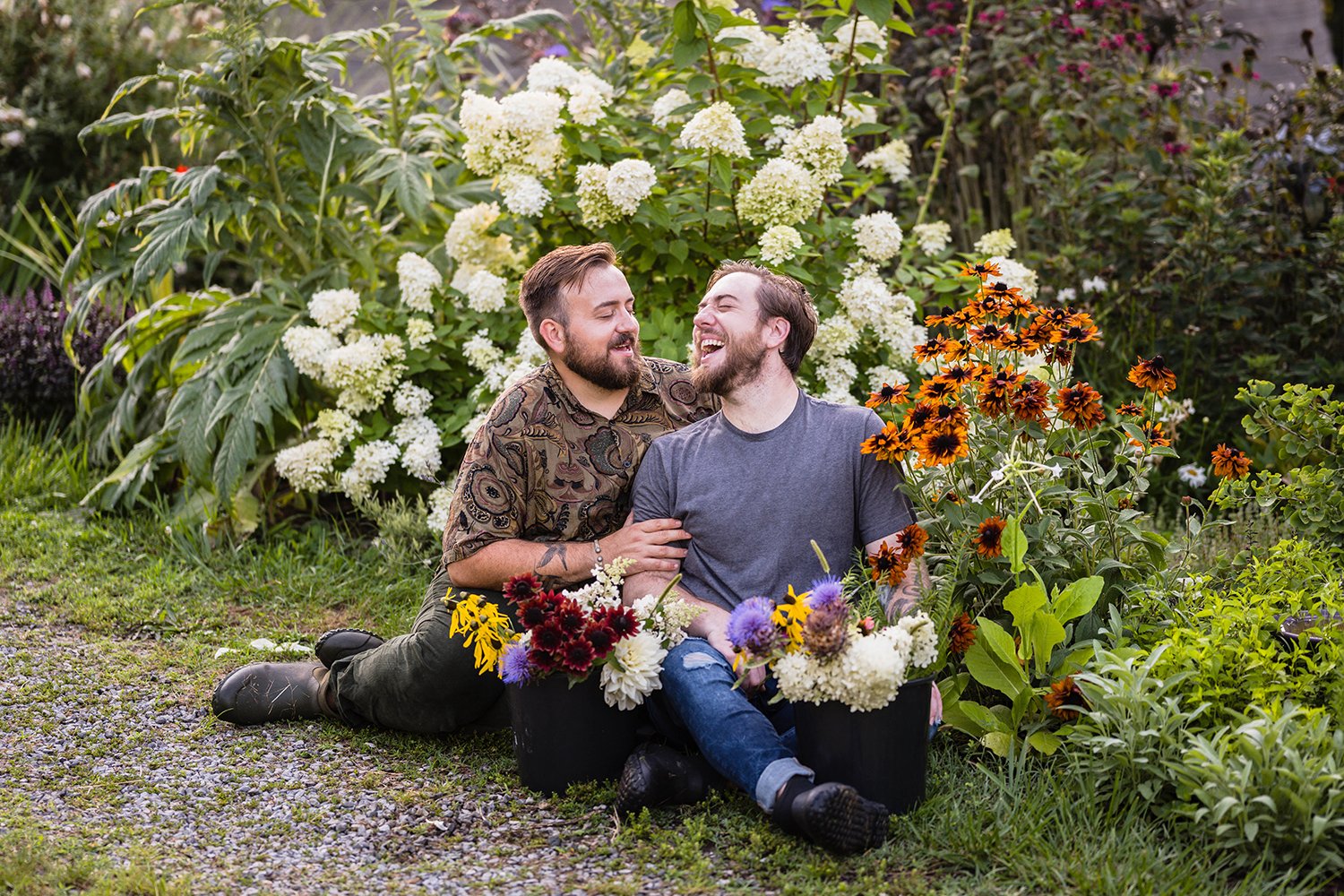 A queer couple sits in their garden at their farm, Fae Cottage Flower Farm with buckets of plucked flowers near them. The couple laughs as they look at one another during their couple’s session.