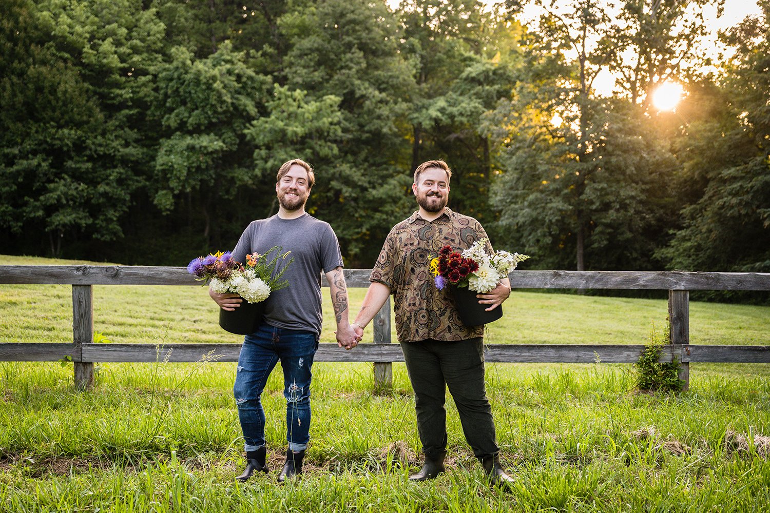 Two gay men hold hands and hold onto buckets with Fae Cottage Flower Farm flowers at their hip and smile towards the camera during their sunrise adventure session.