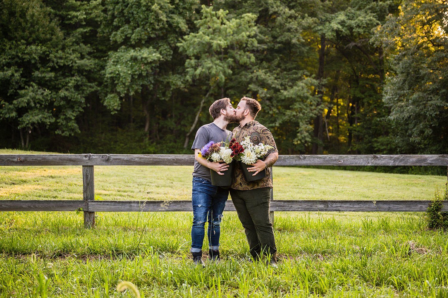 Two queer men hold onto buckets of Fae Cottage Flower Farm flowers at their hip and kiss one another during their sunrise adventure session.