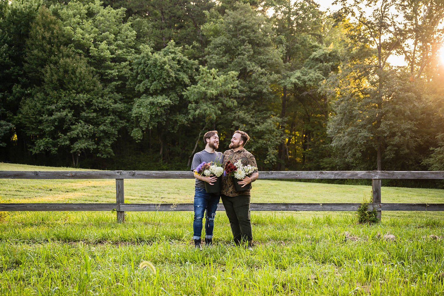 A gay couple holds buckets of flowers in their farm, Fae Cottage Flower Farm. They embrace and look at one another lovingly.