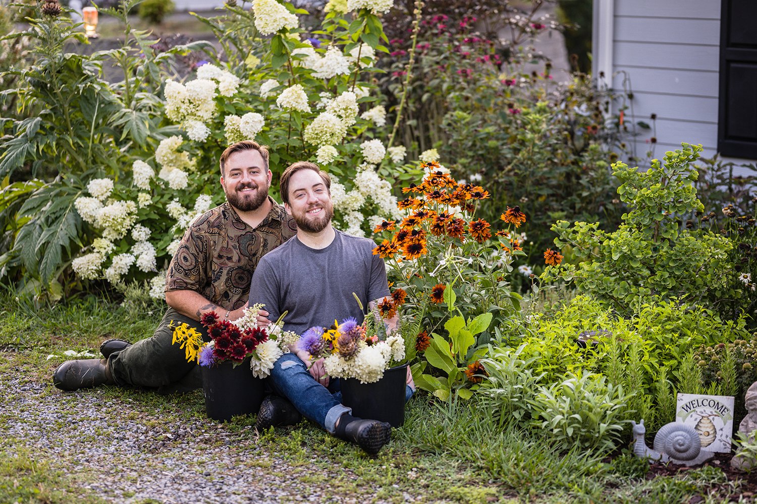 A gay couple sits in their garden at their farm, Fae Cottage Flower Farm with buckets of plucked flowers near them. The pair smile towards the camera during their photoshoot.