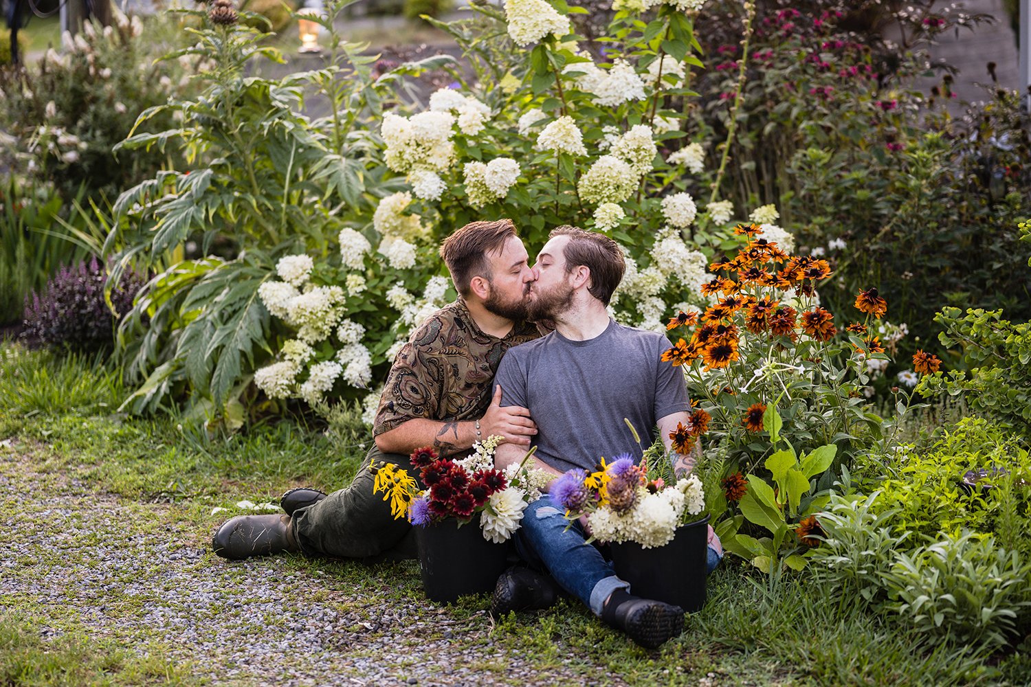 A queer couple sits in their garden at their farm, Fae Cottage Flower Farm with buckets of plucked flowers near them. The couple goes in for a kiss during their adventure session.