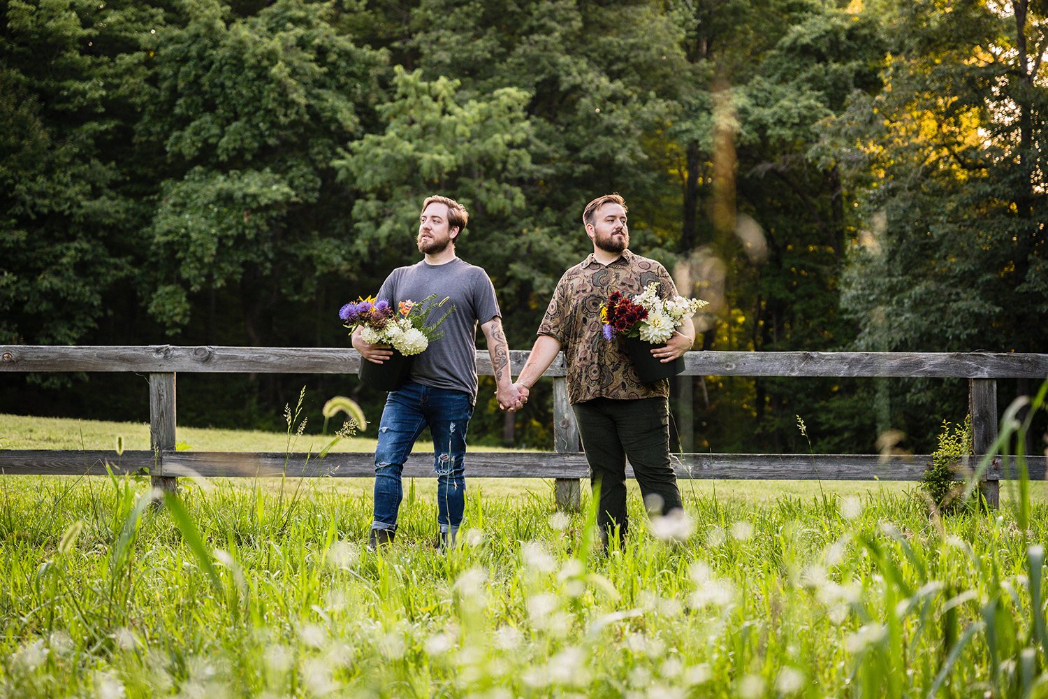 A queer couple holds hands as they hold buckets of flowers at their hip which were plucked from Fae Cottage Flower Farm.