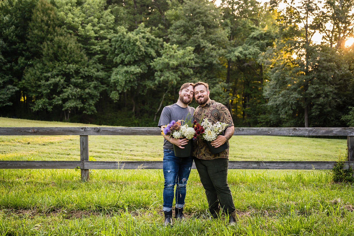 A couple holds onto one another as they show off buckets of flowers from their farm, Fae Cottage Flower Farm, at their hip and smile. 