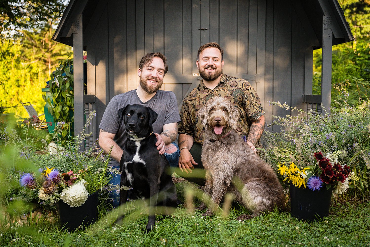 The owners of Fae Cottage Flower Farm poses with their dogs in front of a black shed with buckets of flowers by their side.