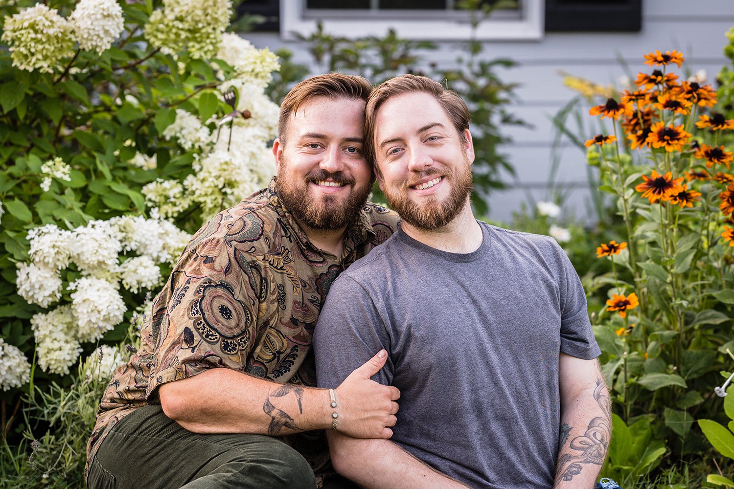 A gay couple sits in front of their garden, which is adorned with flowers, at their farm, Fae Cottage Flower Farm and smiles for the camera during their adventure session.