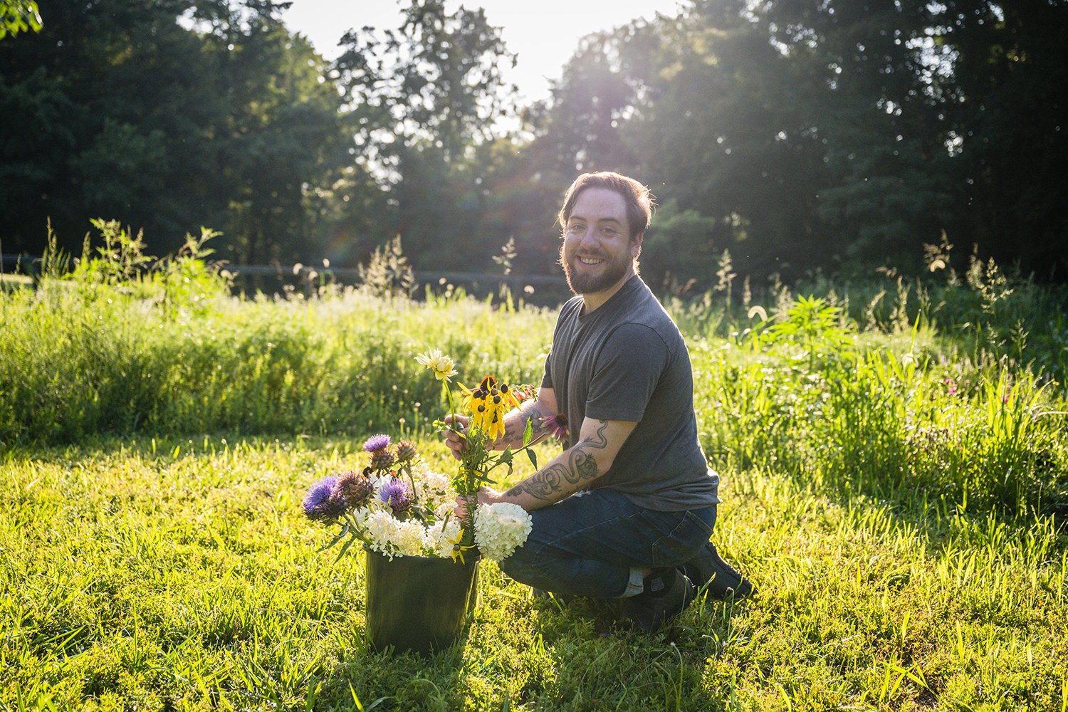 A man crouches and takes some flowers out of a bucket and smiles towards the camera during a photo session at Fae Cottage Flower Farm.