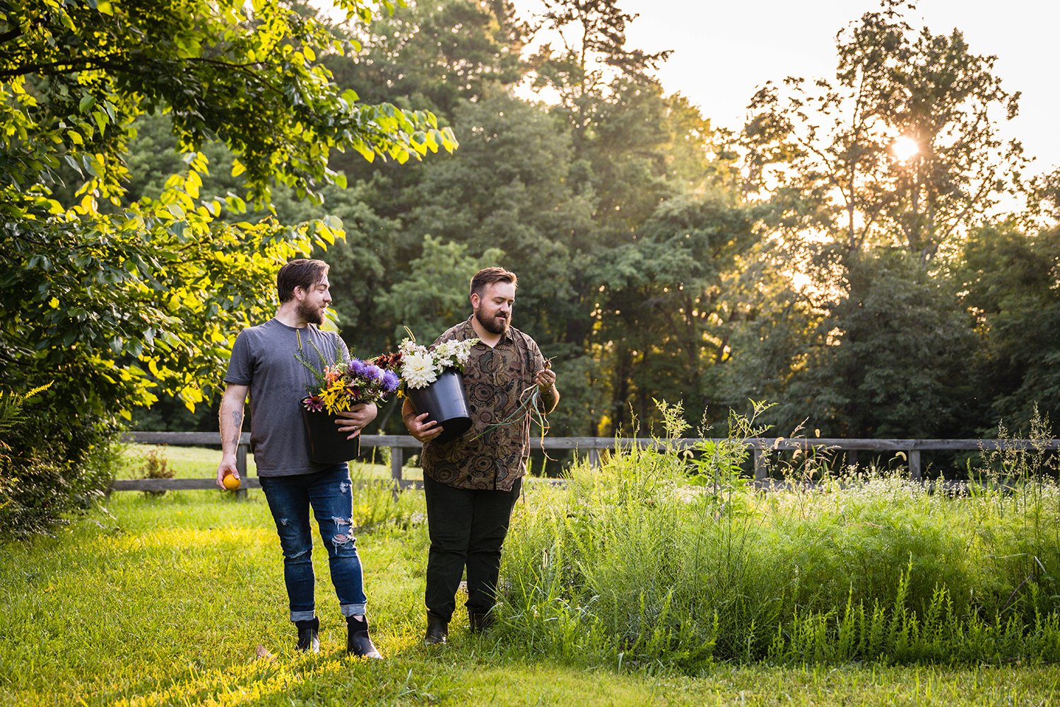 A person holding a bucket of flowers examines another person holding a bucket of flowers and looking at a vegetable they pulled from their farm, Fae Cottage Flower Farm.