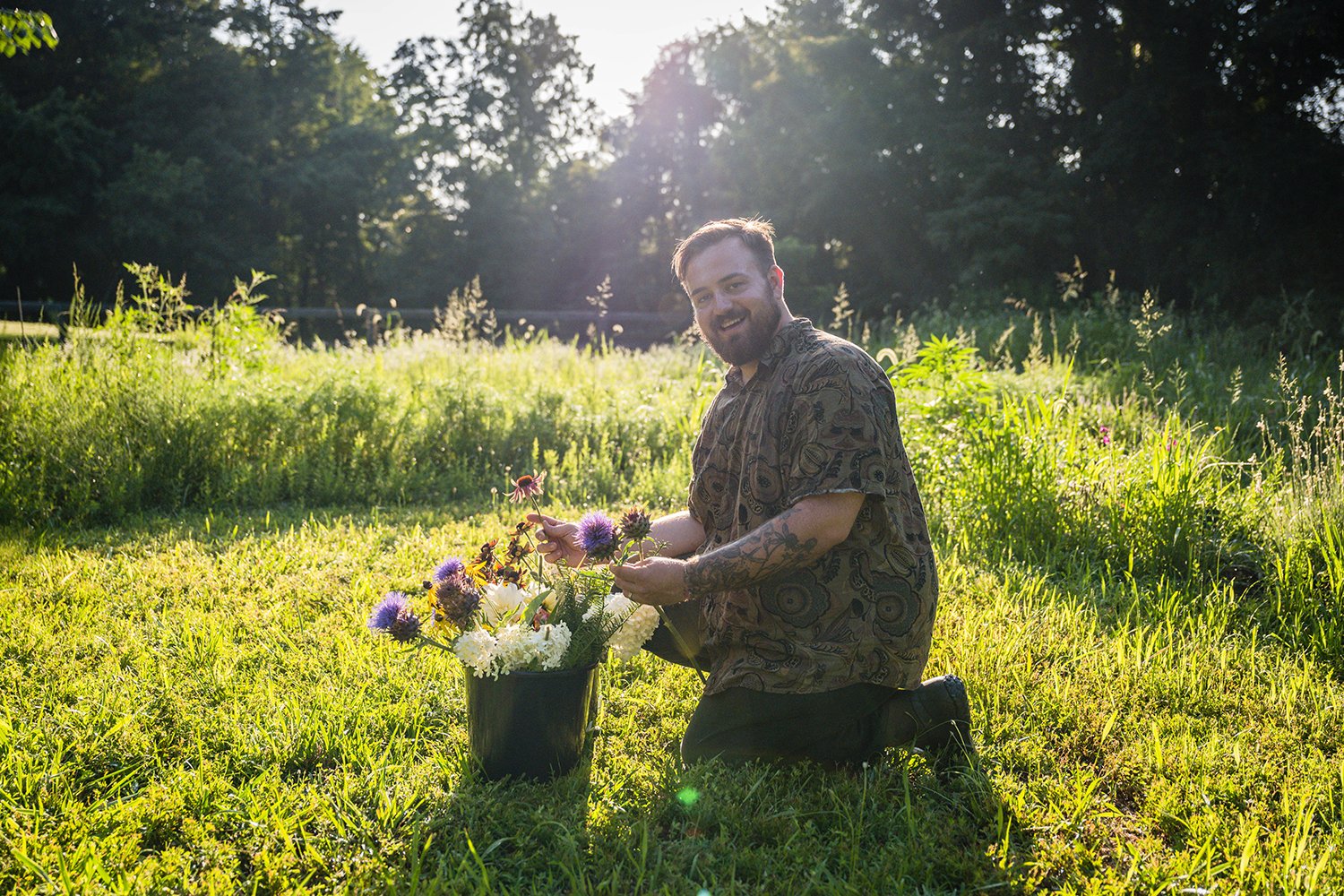 A person crouches and takes some flowers out of a bucket and smiles towards the camera during a photo session at Fae Cottage Flower Farm.
