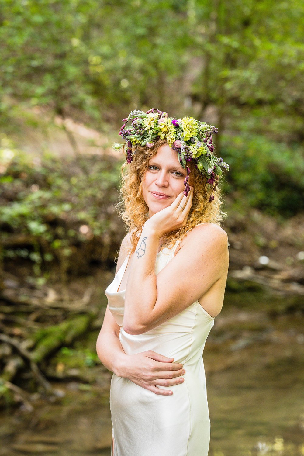 An LGBTQ+ marrier poses wearing a cream satin gown and a flower crown stands in the creek under a canopy of trees for her portraits at Fishburn Park in Roanoke, Virginia.