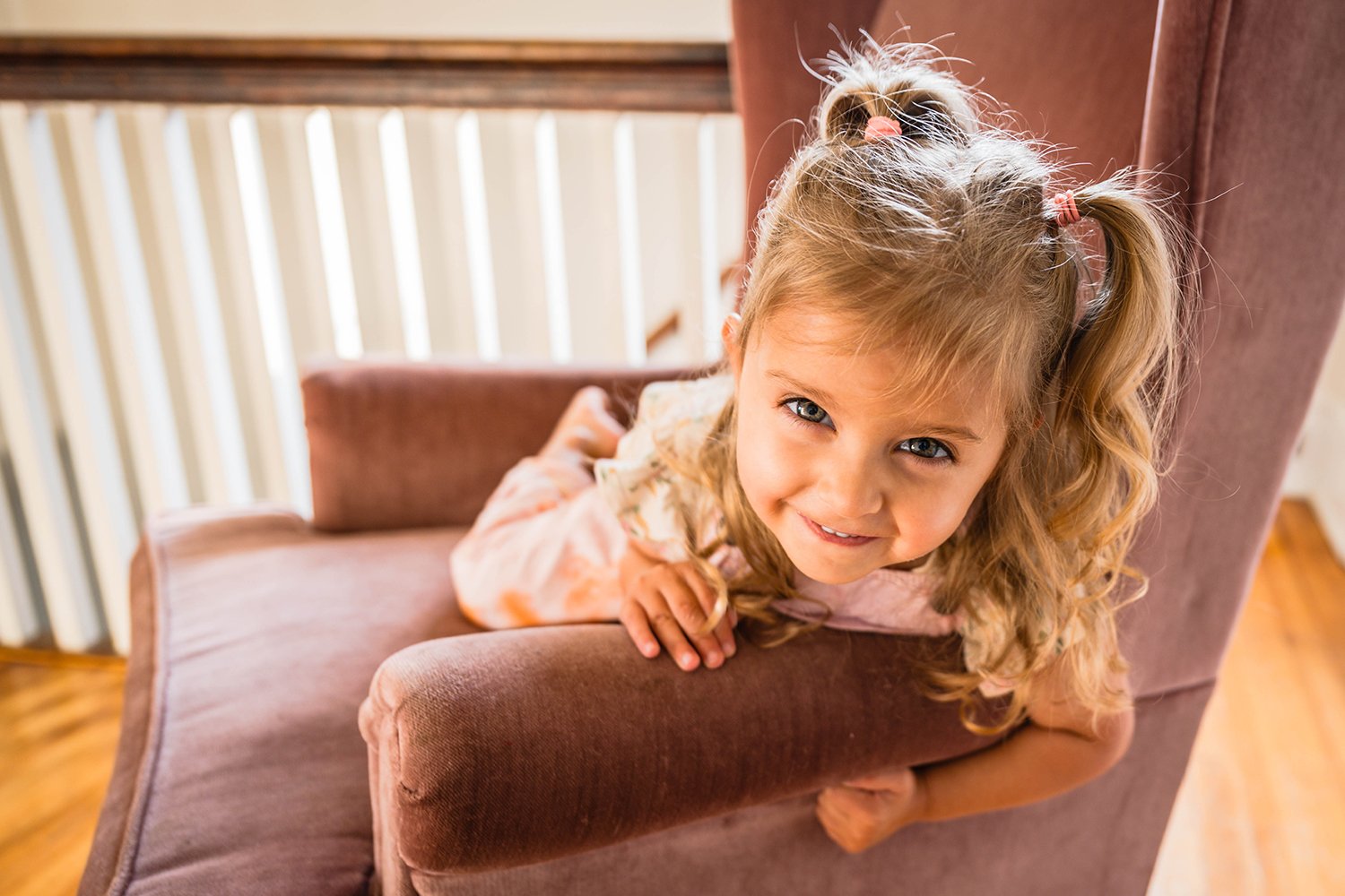 A toddler lays down on a pink chair and hangs off the side to smile towards the camera after getting ready for her mom and dad's elopement day.