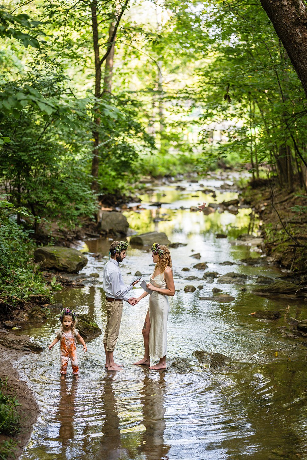 A couple stands in the creek at Fishburn Park to read their vows during their elopement while their toddler stomps around and plays in the water.