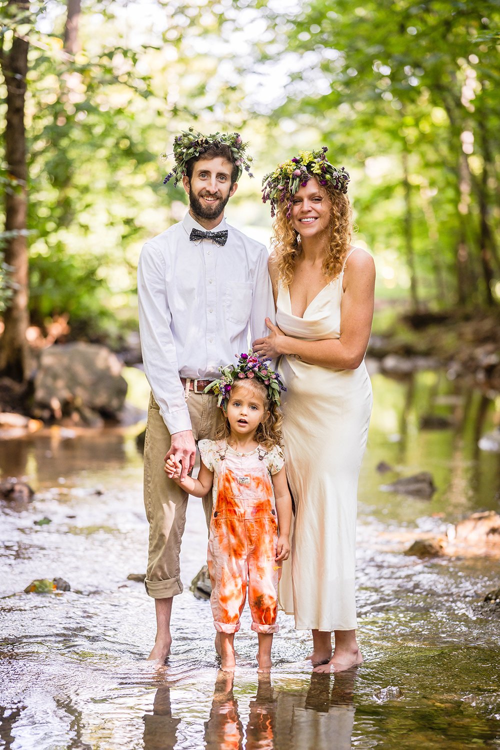 An elopement couple poses with their daughter for a family photo while standing in the creek under a canopy of trees at Fishburn Park in Roanoke, Virginia.