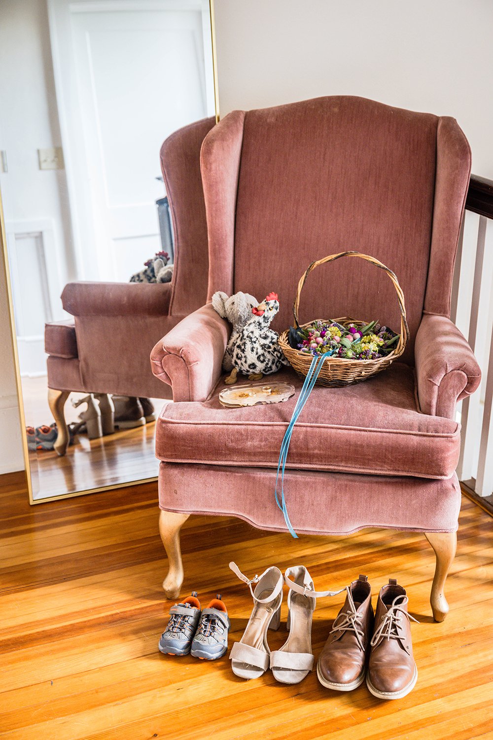 A pink chair in a foyer holds a basket full of flower crowns tied together with blue ribbon and two stuffed animals with three pairs of shoes in front.
