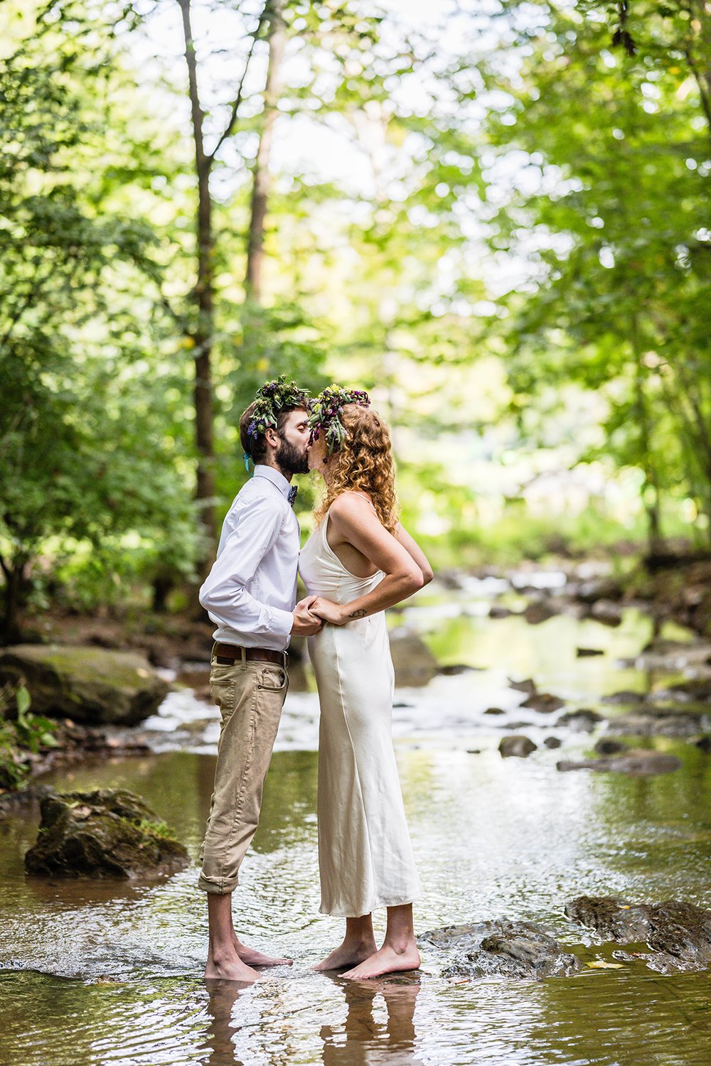 An LGBTQ+ couple kisses after exchanging their rings in a creek under a canopy of trees at Fishburn Park in Roanoke, Virginia during their elopement.