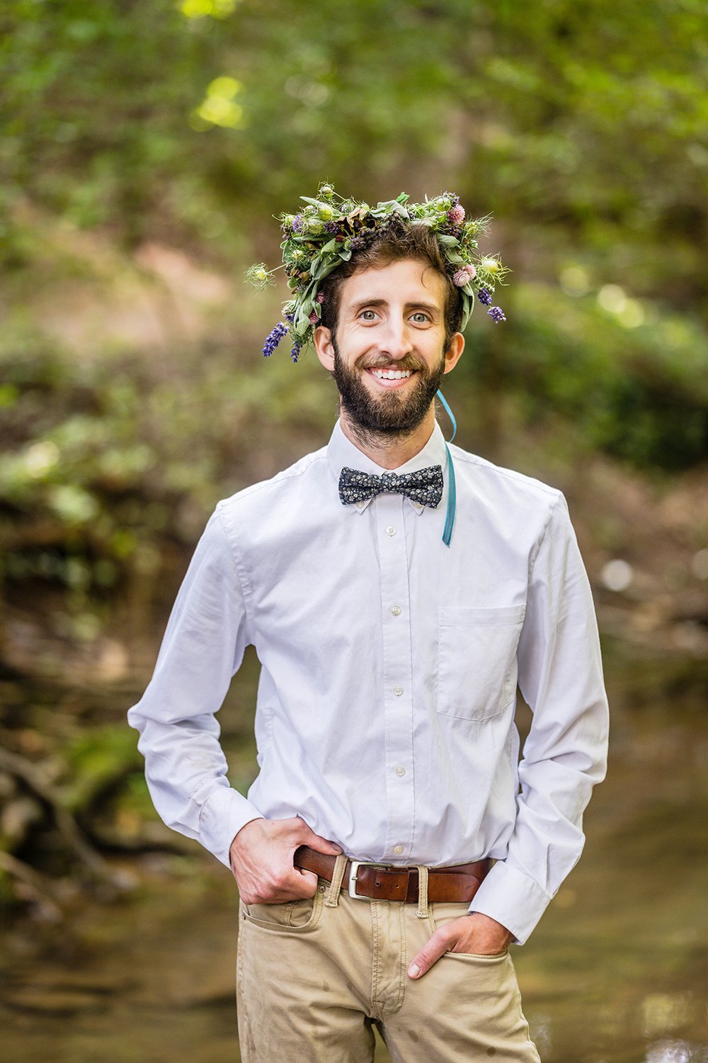 An LGBTQ+ marrier poses wearing a simple button up shirt, bowtie, and khakis and a flower crown stands in the creek under a canopy of trees for his portraits at Fishburn Park in Roanoke, Virginia.