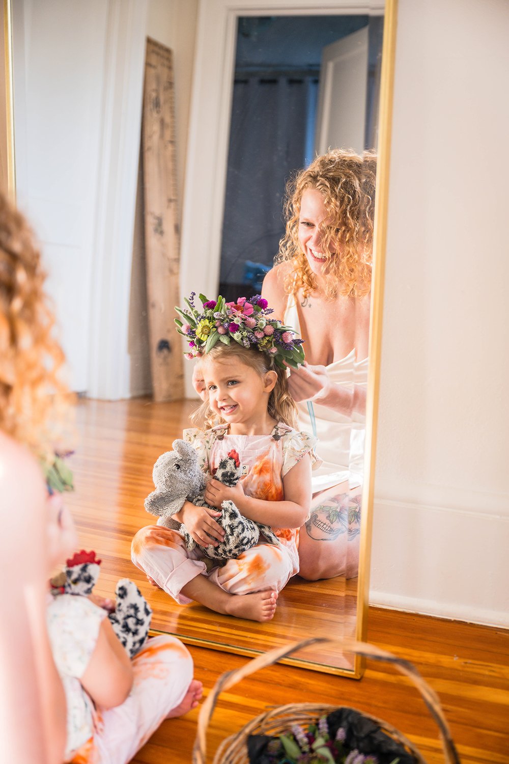 A mother helps put a flower crown on her daughter's head as they both sit in front of a mirror. The daughter smiles and holds two stuffed animals in her lap.