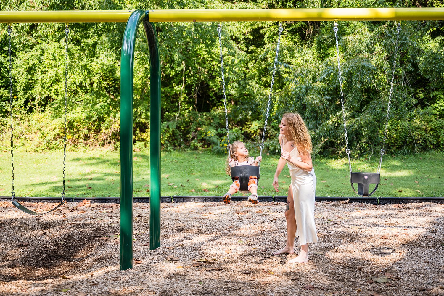 A mother and daughter smile at one another as the daughter swings on a swingset and the mom observes from beside her child.