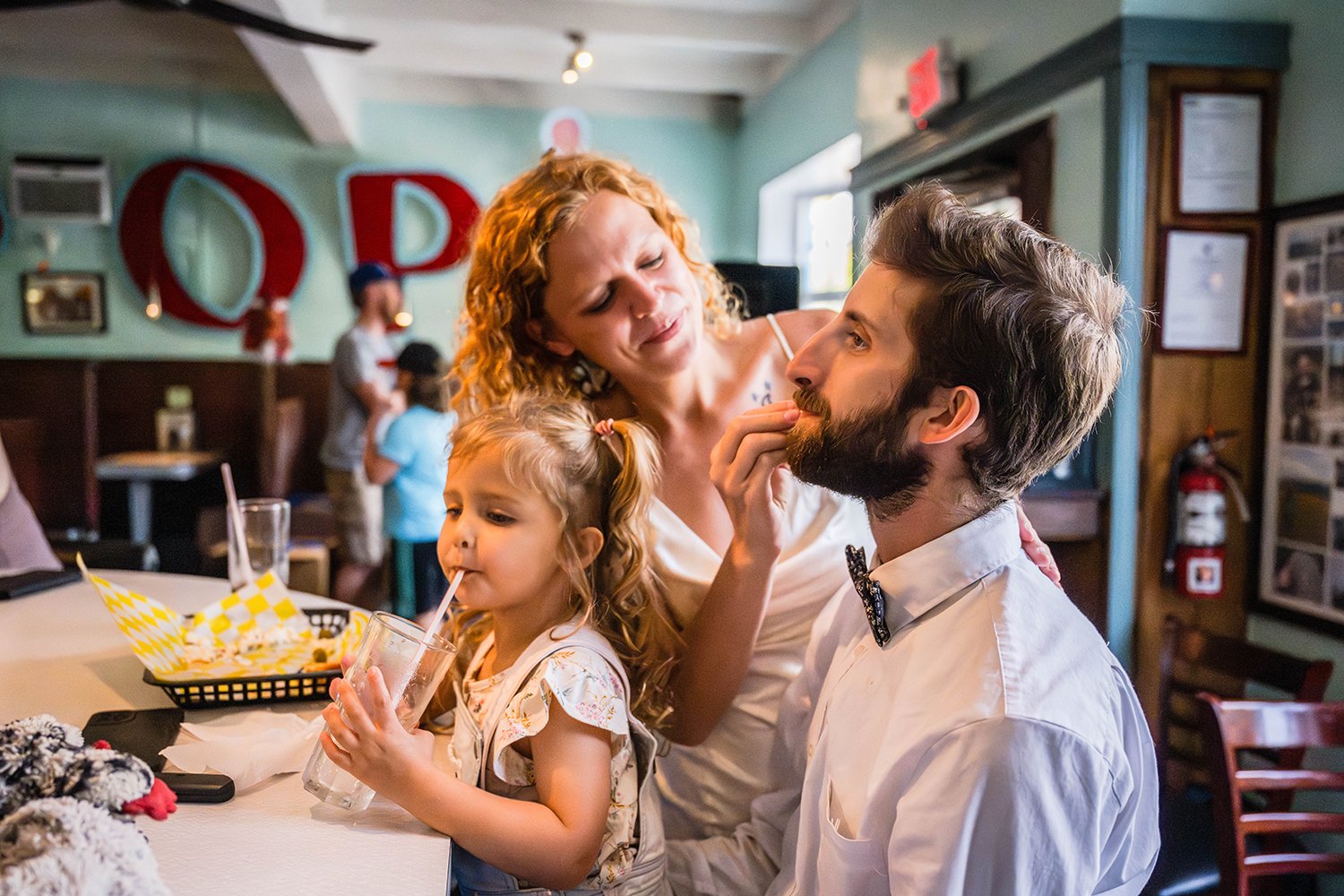 A toddler sits on her father’s lap sipping on a glass of water while her father’s lip is being wiped by her mother inside of Pop’s Ice Cream &amp; Soda Bar.
